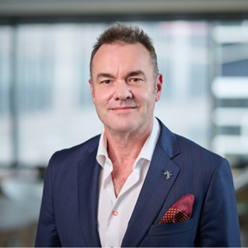 Middle-aged man in navy suit with red pocket square, smiling in office setting.